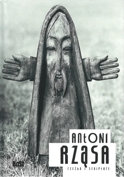 Na okładce widoczna jest drewniana rzeźba stojąca na trawniku przedstawiająca postać skrzata