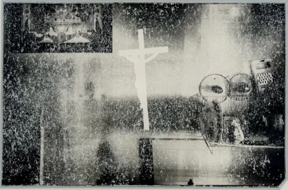 Czarno-biały, surrealistyczny i ziarnisty wydruk przedstawiający duży, abstrakcyjny, biały krzyż umieszczony na teksturowanym, monochromatycznym tle wokół niewyraźnych kształtów i obiektów